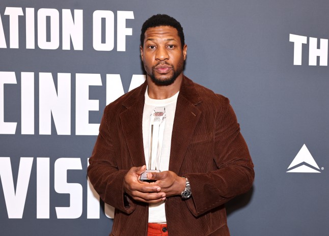 Jonathan Majors at a red carpet event wearing red trousers, a white t-shirt, and a brown jacket