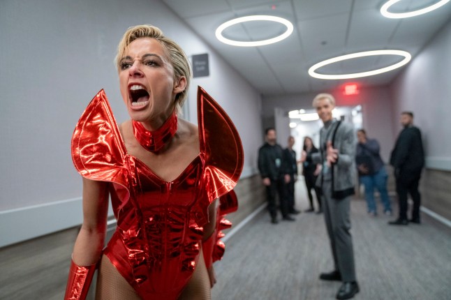 Naomi Scott as Skye Riley in Smile 2. She is wearing a red costume and standing in a corridor of people screaming