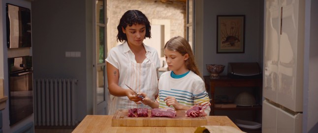 Carla Diaz as Teodora and Naila Schuberth as Alba cut meat together in the kitchen in Delicious
