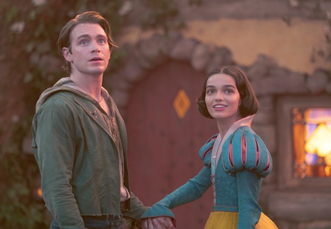 Andrew Burnap as Jonathan and Rachel Zegler as Snow White hold hands as they look at fireflies in front of the dwarfs' cottage in Snow White