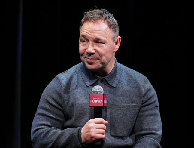 Stephen Graham speaks onstage holding a microphone while wearing a grey knitted top