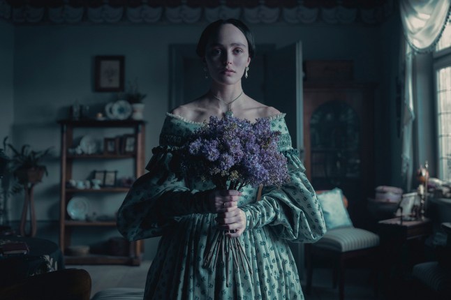 Lily-Rose Depp as Ellen Hutter, holding a bouquet of purple flowers and standing in a parlour, in a scene from Nosferatu