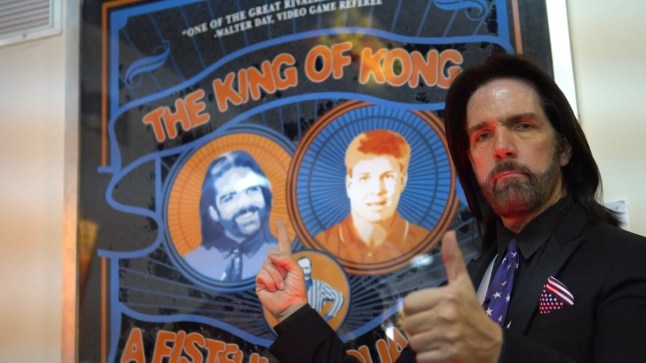 Billy Mitchell giving a thumbs up in front of a poster for The King of Kong documentary