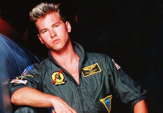 Val Kilmer in his Top Gun role, wearing the green flying suit and leaning on his knee.