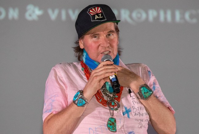 Val Kilmer speaks at a Top Gun screening in 2019, wearing a pink shirt, turquoise jewellery and a black baseball cap