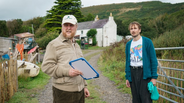 Tim Key as Charles carrying a whiteboard and Tom Basden as Herb McGwyer with a plastic bag, standing behind him by a gate in The Ballad of Wallis Island