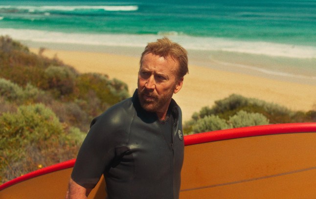 Nicolas Cage in The Surfer, holding a surfboard with the beach in the background