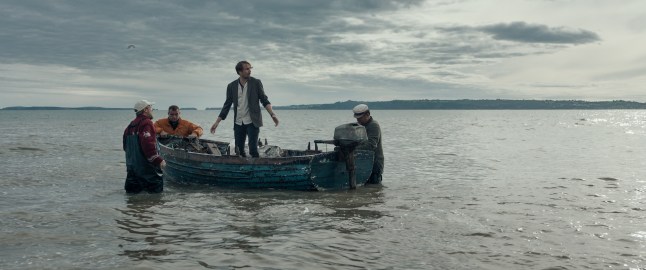 Tom Basden as Herb McGwyer stands awkwardly in a rowing boat by the shore in The Ballad of Wallis Island