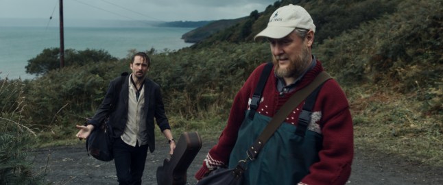 Tom Basden as Herb McGwyer carries a guitar case up the hill, wet, following exasperatedly behind Tim Key as Charles Heath in a baseball cap, jumper and overalls in The Ballad of Wallis Island