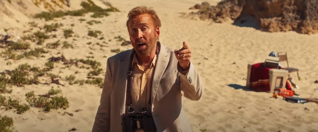 Nicolas Cage in a dirty linen suit pointing his finger and looking wild and unkempt on the beach in a scene from The Surfer