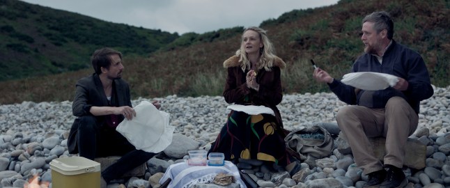 Tom Basden as Herb McGwyer, Crey Mulligan as Nell Mortimer and Tim Key as Charles Heath sit around a fire on a stony beach in The Ballad of Wallis Island
